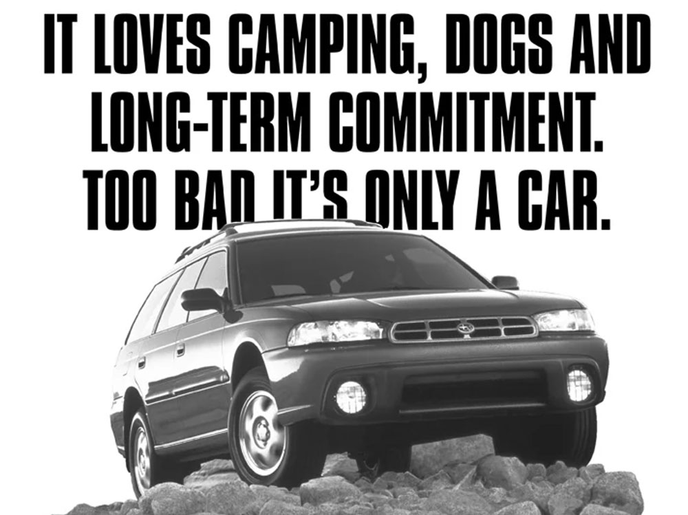 A black and white Subaru ad from the mid ‘90s, showing a dark-colored Subaru crossover vehicle parked on top of a rocky hill. Above and copy behind it, in big black letters, reads, “It loves camping, dogs, and long-term commitment. Too bad it’s only a car.”