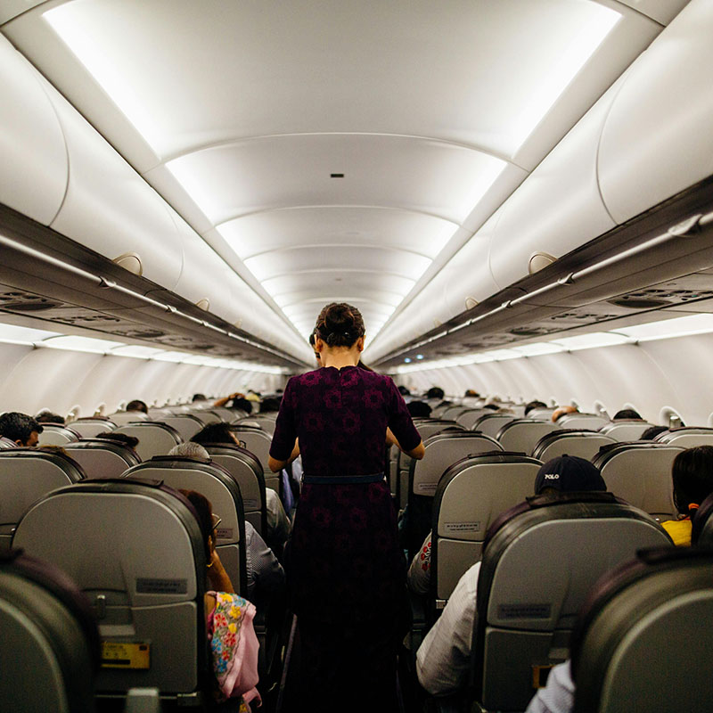 Photo looking down the aisle of an airplane from the back, showing rows and rows of gray seats with the tops of heads poking up. Standing in the aisle several feet away is a flight attendant, wearing a burgundy dress, with her dark hair in a bun, facing away from us and looking slightly downward as if at a drink cart.
