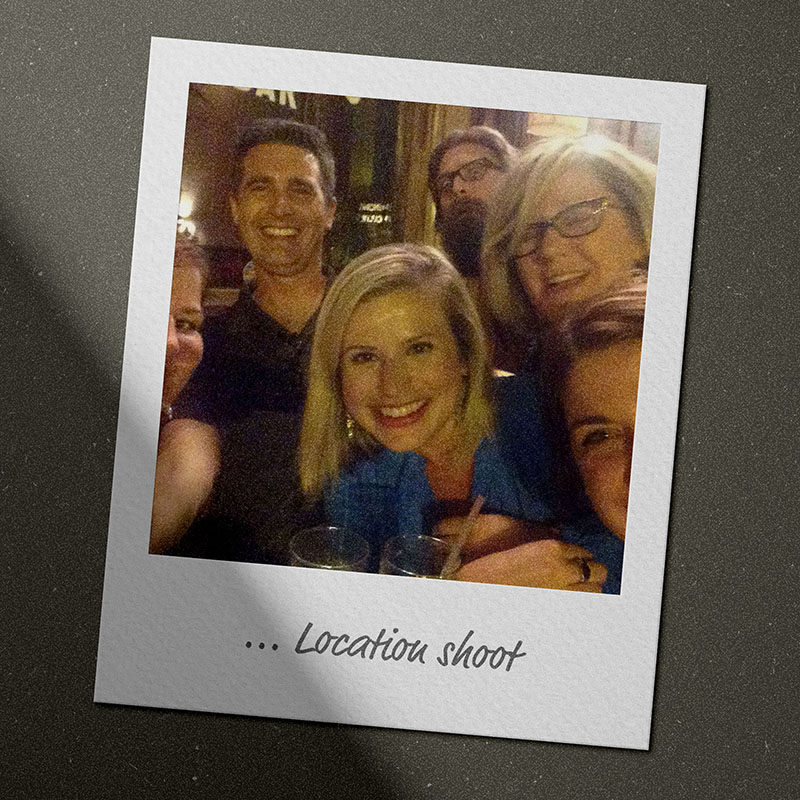 White-edged Polaroid photo mockup lying on a dark gray surface. The photo shows a hazy image of six people crowded into a nighttime selfie in front of a bar, including a young woman with a blonde bob and a blue button-down shirt, a man with dark hair and a black polo shirt, a middle-aged woman with a blonde bob, and the edges of two women’s heads just peeking in at opposite corners. And a photobomb of a guy with brown hair, glasses, and a long brown beard. The caption in handwritten font at the bottom of the photo reads, “… Location shoot.”