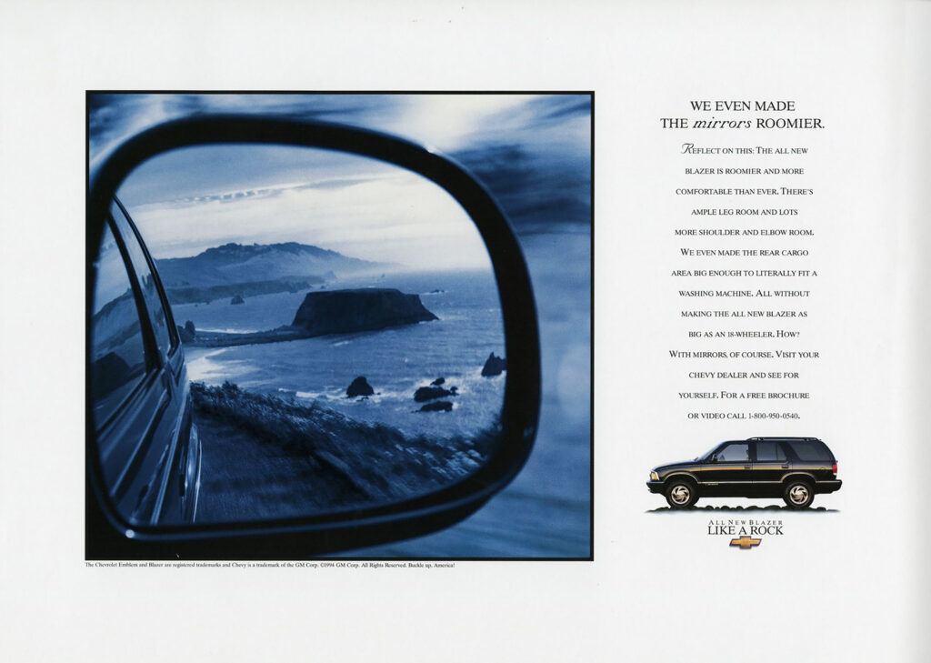 A large, blue-toned black-and-white close-up photo of a car wing mirror showing a bayside-looking image with a large rock in the middle of the water and hills and cliffs in the background. To the right of the photo are the words: “We even made the mirrors roomier. Reflect on this: The all-new Blazer is roomier and more comfortable than ever. There’s ample leg room and lots more shoulder and elbow room. We even made the rear cargo area big enough to literally fit a washing machine. All without making the new Blazer as big as an 18-wheeler. How? With mirrors, of course. Visit your Chevy dealer and see for yourself. For a free brochure or video call 1-800-950-0540.” Below that is a small side-view photo of a black Chevy Blazer. Underneath it, “All New Blazer. Like a Rock” and the Chevy logo.