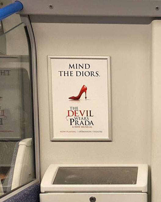 Small poster on the wall of a London tube car. The white poster shows the words “Mind the Diors” above a side shot of an extremely high red stiletto shoe with a pointy devil tail at the point of the heel. Below it are the words, “The Devil Wears Prada, a New Musical. Now playing, Dominion Theatre.”
