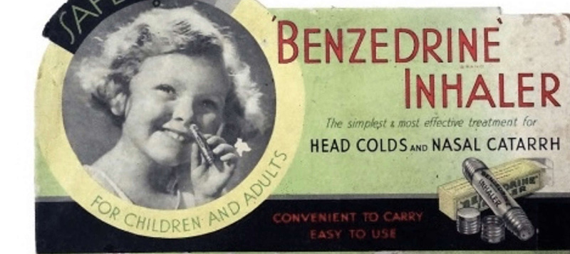 A very old ad with a green background shows a circular black-and-white image of a young, smiling child with a tube up to her nose. Around the picture is a circle reading, “Safe for children and adults,” The headline in red text reads, “‘Benzedrine Inhaler’,” and under it is smaller text reading, “The simplest & most effective treatment for head colds and nasal catarrh.” At the bottom, in red letters on a black stripe, are the words, “Convenient to carry, easy to use,” Followed by the image of a silver Benzedrine inhaler and a few accessories I can’t identify.