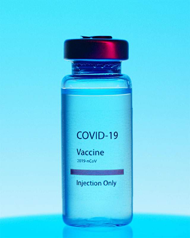 A glass vial labeled “COVID-19 Vaccine, 2019-nCoV, Injection Only,” filled with clear liquid, sits on a glowing turquoise-blue surface against a turquoise-blue background.