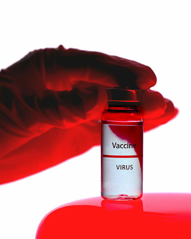 A rubber-gloved hand, lit from the bottom in eerie red light, touches the top of a glass vial labeled “Vaccine Virus” filled with clear liquid, sitting on a red glowing surface against a white
