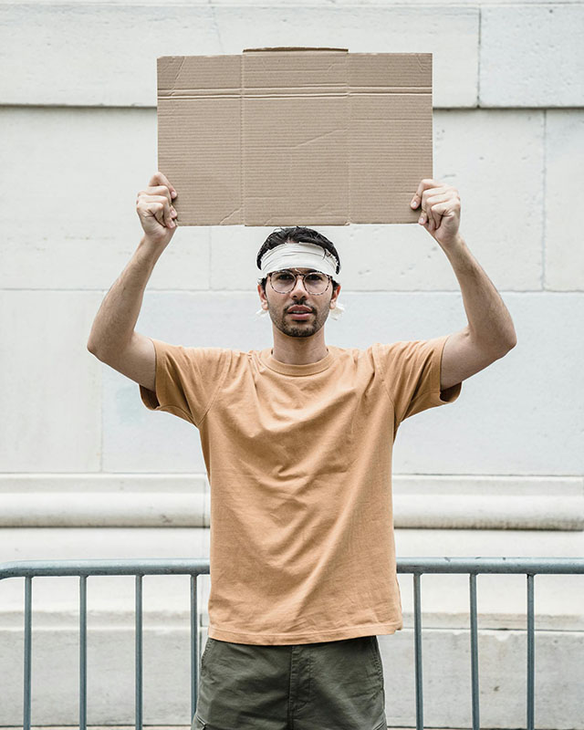 A young, dark-haired man in a faded orange t-shirt and army-green pants, with a strip of white fabric around his head above his eyes, with glasses and a scruffy beard, stands in front of a green barrier fence with a white concrete-block wall in the background. He’s holding a cardboard sign above his head, but it’s blank.
