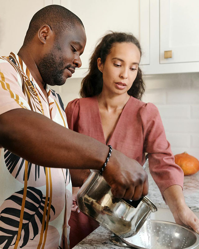 A man and a woman are cooking together in an all-white kitchen. He’s tall and heavyset, wearing a Hawaiian shirt in muted tones, and has close-trimmed hair and a beard. She has long dark hair, held in a black headband, and is wearing a pretty rose-colored linen v-neck dress. He’s pouring water from a sauce pot into a metal colander, and she’s holding the colander. Their gazes are both directed at the pouring water.