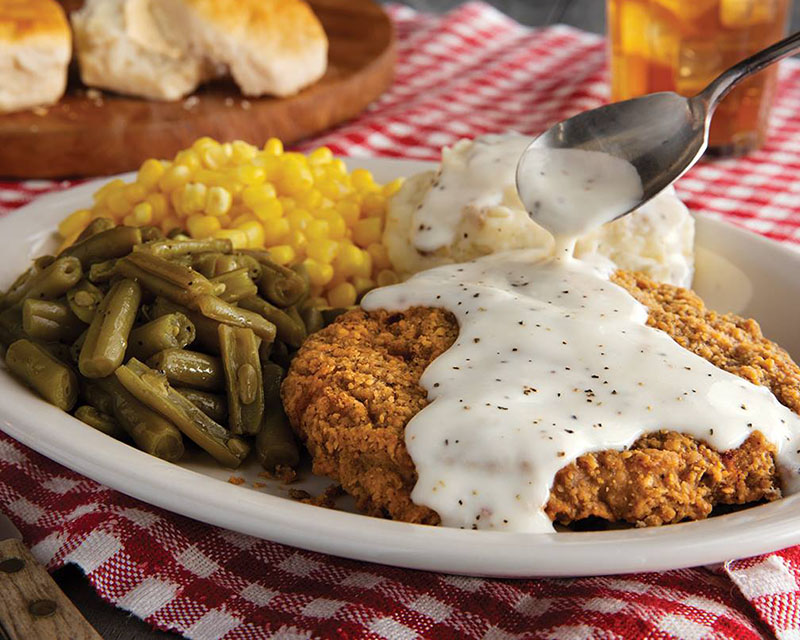 A photo of a chicken dinner from Cracker Barrel, sitting on a red gingham tablecloth with a plate of biscuits and a glass of tea in the background. The white dinner plate features a piece of fried chicken smothered in gravy, with a spoon above it dripping gravy onto it. Clockwise from the left are a pile of green beans, a pile of corn, and and a serving of mashed potatoes with yet more gravy on it.
