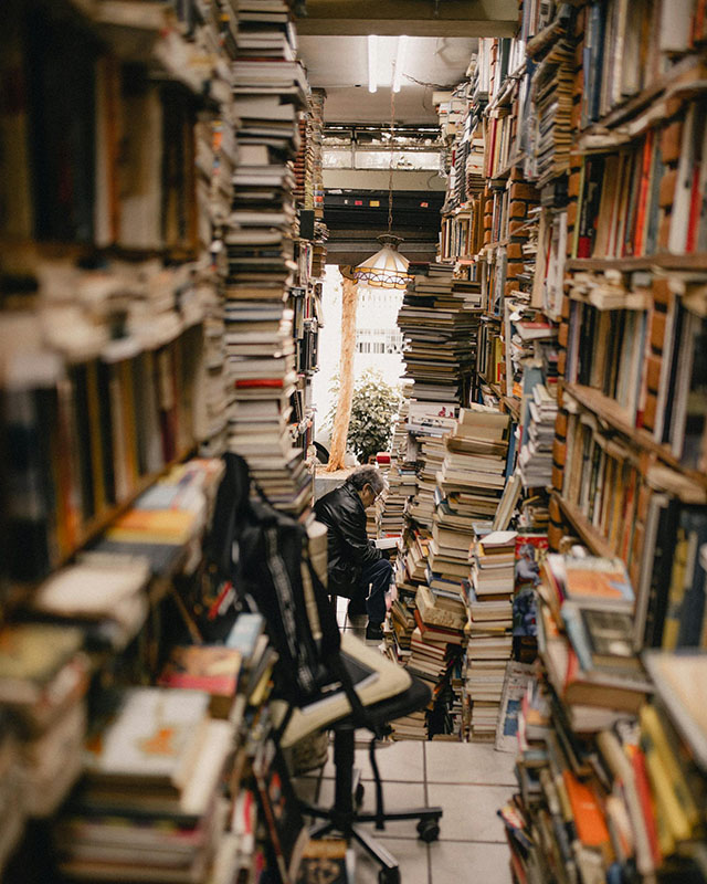 A photo of an office-like space, cramped and narrow but long, with bookcases on both sides that are bursting with books. The floor is also covered with tall stacks of books, and a man in dark clothes is sitting toward the other end of the space, tiny, reading a book.