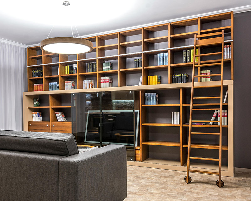 Photo of a large, light-wood bookcase with an open back, spanning the width of a gray-pained room, with a gray sofa sitting in front of it and a window covered with sheer curtains to our left. The bookcase has a ladder and a few sets of books on different shelves, but for the most part, it’s empty.