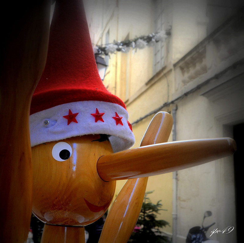 A close-up of a wooden Pinocchio doll, so all you can see are his eyes, his long nose, his red smile, and and his hat, which is red and pointed with a white brim with red stars on it. The background, just out of focus, appears to be the graying front of a row of white townhouses.