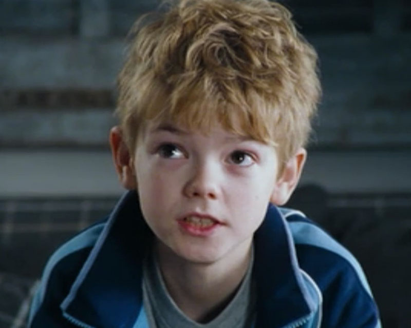 A cropped screenshot from the move “Love, Actually,” showing a young boy sitting on a bench in a blue zip-neck jacket and a blue t-shirt. He has curly blonde hair and adorable dark eyes, and he’s gazing up at something out of frame to the left with a kind of sad yet hopeful expression that would make you give him anything.
