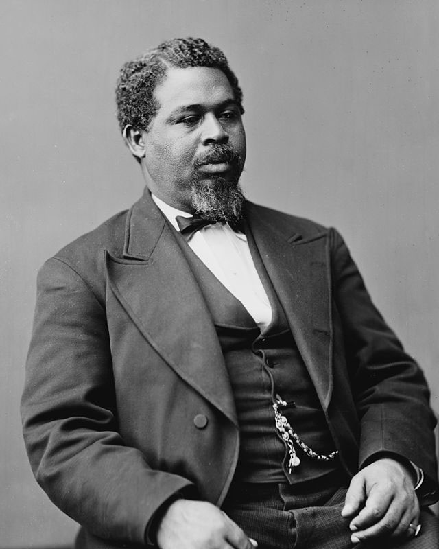 Black-and-white photo portrait of U.S. Representative Robert Smalls, circa the 1870s. He’s a black man with curly, graying black hair and a graying black goatee. He’s sitting tall, his gaze toward someone to the left of the frame, and he’s wearing a black suit jacket, dark gray trousers with a faint check, a black waistcoat, a white shirt, and a black bowtie. He has what I’m assuming is a gold watch chain hanging from a buttonhole to a watch pocket hidden under his jacket, and a ring on his left pinky finger.