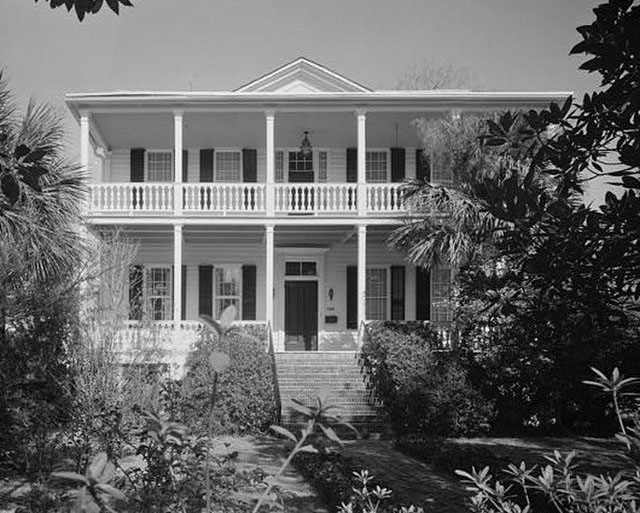 Black and white photo of a white plantation house with black shutters, two stories with white columns and porticos on both floors. It has a large bush on either side of the stairs to the front porch, and large magnolia and palmetto trees on either side of the brick path to the steps.