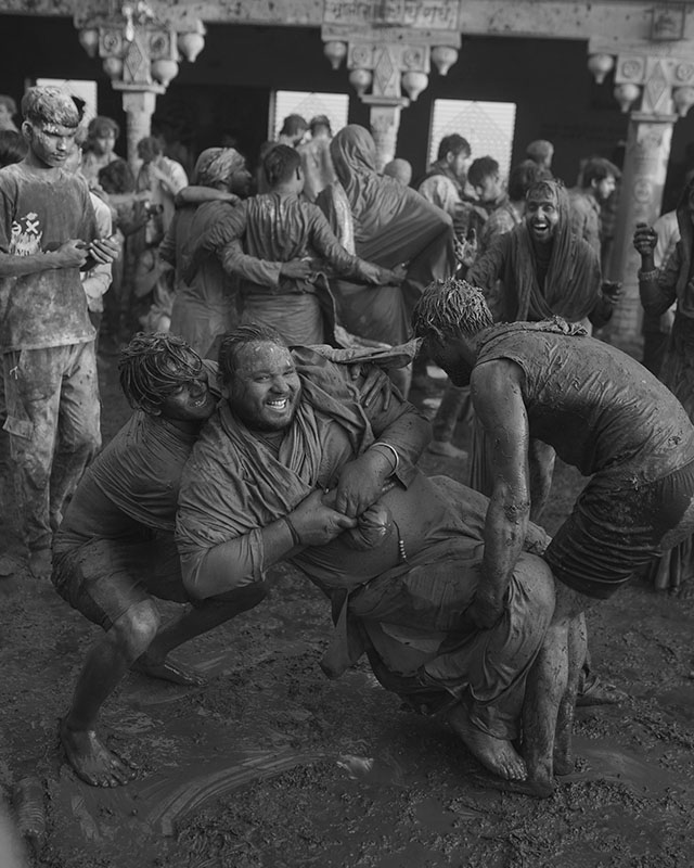 Black-and-white photo of three men wrestling, smiling and completely covered, every inch of them, in mud. A dense crowd behind them, also muddy, mostly doesn’t even notice, with their backs turned. The crowd is standing under what appears to be a stone portico held up by elaborate, very aged columns.