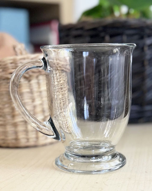A close-up photo of a clear glass cafe mug sitting on a light-wood coffee table. In the background are, on the right, a lily planted in a dark woven planter and, on the left, the side of a lidded container made of woven pale reeds.