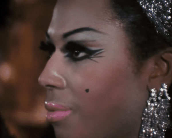 Close-up photo of Black drag queen Crystal LaBeija, taken at the Miss All-America Camp Beauty Pageant in 1967. We can see her heavy, black, winged eyeliner, pink lips, arched eyebrow, and heart-shaped beauty mark on her cheek, as well as her ear with two large, dangling crystal earrings and the front of her hair with a matching crystal ornament.