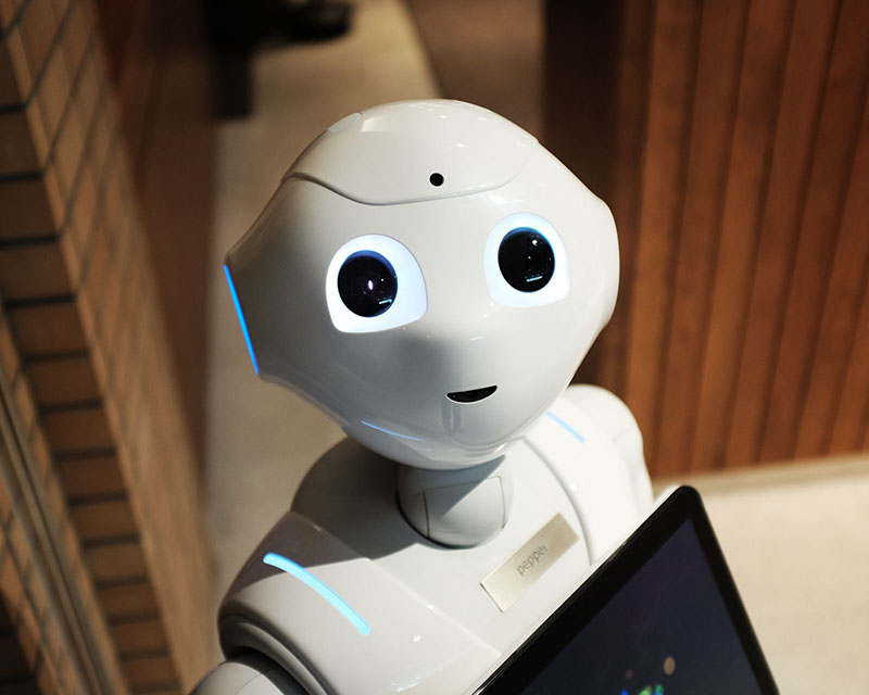 Close-up photo of a cute, plastic white robot, standing in what appears to be a beige-carpeted room with natural wood-paneled walls. The robot has a little round head, big lit-up eyes, blue-lighted accents on its ears and shoulders, and a black iPad attached to its chest, as well as a brass nameplate that reads “Pepper.”