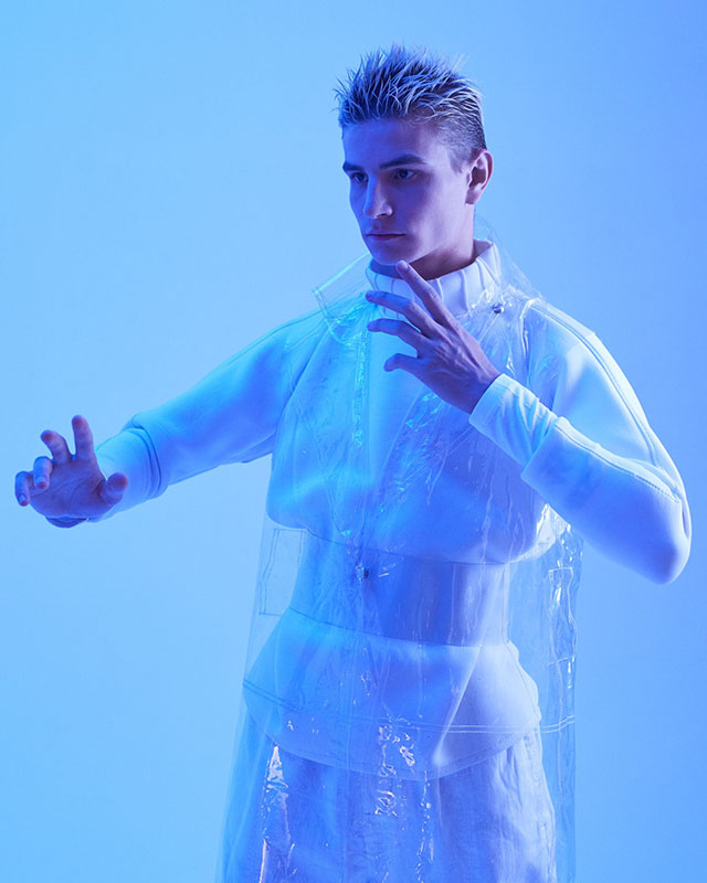 Photo of a man from above the knees up. He has spiky, bleached blonde hair and is swearing what looks like a clear cellphone poncho over a white neoprene long-sleeved crop top and shorts, and he’s lit with blue light, standing against a light-blue wall. He’s standing frozen with his arms half-extended in front of him, much like he’s pretending to be a robot (or possibly doing The Robot).