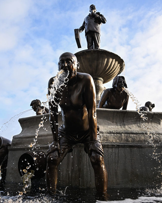 A photo of Channel 4’s “Fountain of Filth” installation, shot under a blue sky from a low angle to show a bronze figure of a man hunched over and appearing to vomit brownish water in front of a large, stone pedestal, with other figures on and around the pedestal doing the same thing. At the very top of the fountain is the bronze figure of an executive, holding his lapel, his pockets and briefcase bulging with cash.