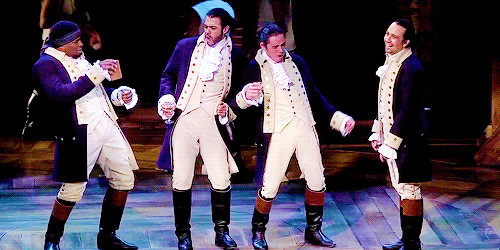 A gif from the Broadway musical “Hamilton,” showing four Black and Latino men in colonial military garb — high black boots, buff breeches, buff waistcoats, navy military jackets with brass buttons, and ruffled white cravats — standing in the middle of a stage. Three of them are dancing salaciously at the fourth, who is turning away and laughing.