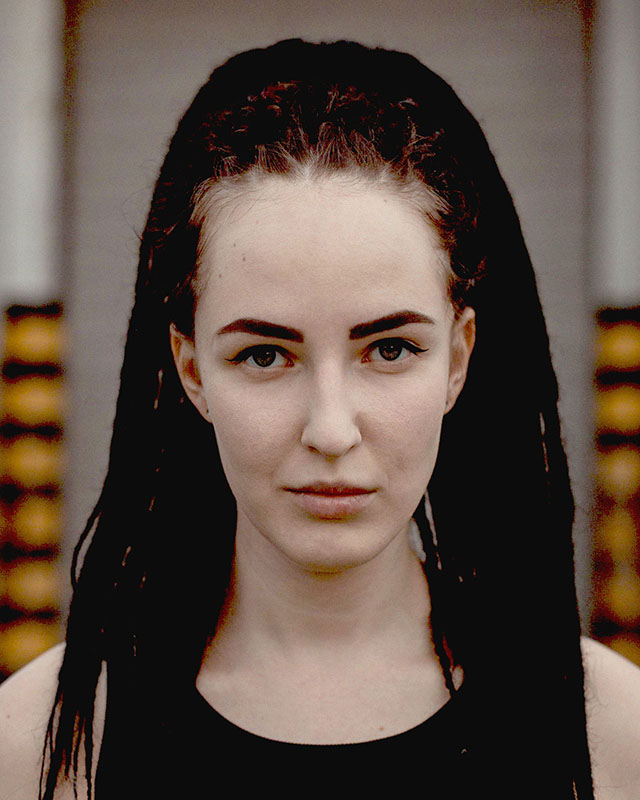 Headshot of a woman against a vaguely urban/industrial background. She’s a white woman staring directly at the camera, with long, reddish hair done up in braids and dreadlocks, minimal makeup, and a black tank top.