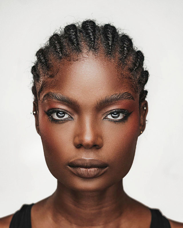Headshot of a woman against a white background. She’s a medium-skinned Black woman with light eyes and fantastic cheekbones, staring boldly at the camera, wearing a black tank top, with dramatic eyeliner and tidy cornrows.