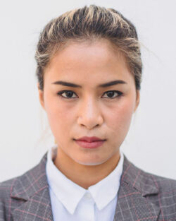Headshot of a woman against a pale gray background. She’s a young Asian woman with minimal makeup and blonde-streaked hair brushed smoothly back into a tidy hairdo, staring straight at the camera, and she’s wearing a white blouse buttoned all the way up and a gray plaid blazer.