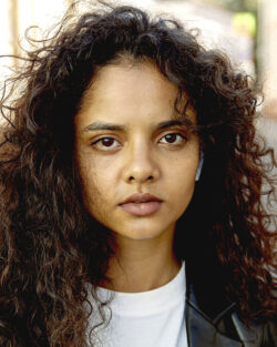 Headshot of a woman against a vaguely leafy background. She’s a young, light-skinned black woman with minimal makeup and cascades of curly, dark hair, staring straight at the camera, wearing a white t-shirt and black leather jacket.