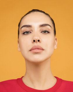 Headshot of a woman against a yellow-orange background. She’s a young woman with closely cropped hair and minimal makeup, staring defiantly at the camera, wearing a red t-shirt.