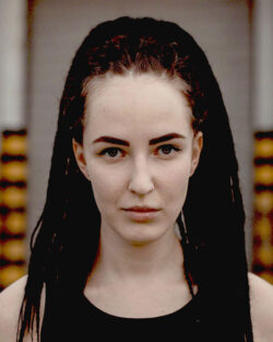 Headshot of a woman against a vaguely urban/industrial background. She’s a white woman staring directly at the camera, with long, reddish hair done up in braids and dreadlocks, minimal makeup, and a black tank top.