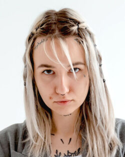 Headshot of a woman against a white background. She’s a white woman staring grimly at the camera, with stringy, blonde, shoulder-length hair, with some of it trailing into her eyes, Her hair is long, below her shoulders, but parts of the front are braided and pinned back enough to show the tattoos running along her hairline, under her left eye, under her chin, and across her upper chest and collarbones, visible between the lapels of her roomy gray blazer.
