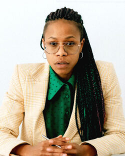Photo of a woman seated at a table with her hands folded in front of her, looking intently at the camera, against a white background. She’s a medium-skinned Black woman with long, black braids pulled into a ponytail draping over her shoulder, with round wire-rimmed glasses, a green button-down blouse with tiny black polka dots, and a light-yellow windowpane-check blazer with strong shoulders, and she has a silver pen woven between her fingers.